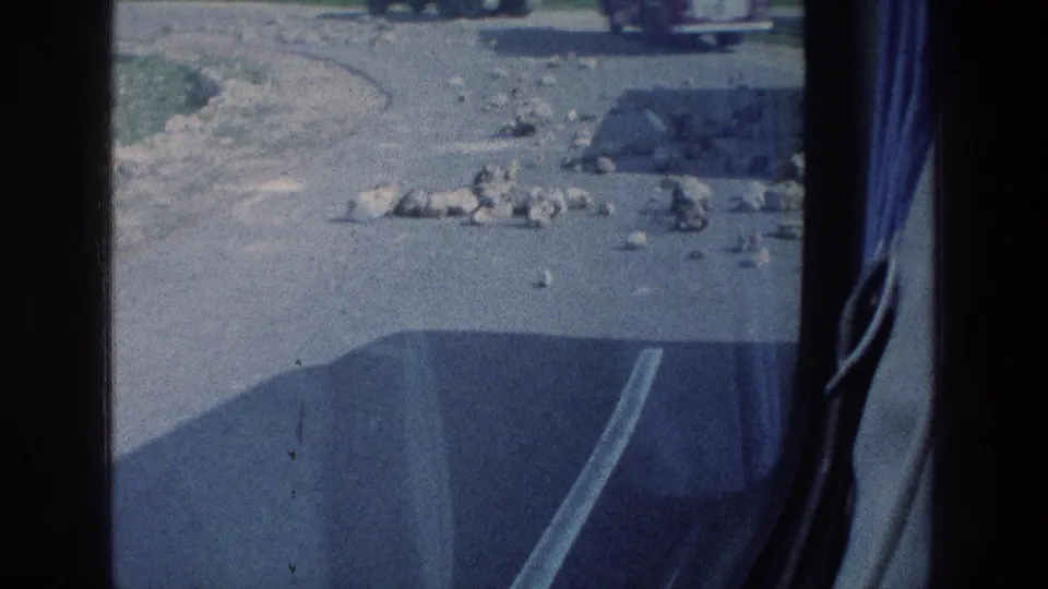 1976 8mm travel view from inside a vehicle, showing scattered stones on a paved road and distant vehicles beyond the window frame.