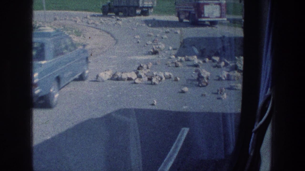 Vehicle-window view of rocks on a road with trucks or cars in the distance.