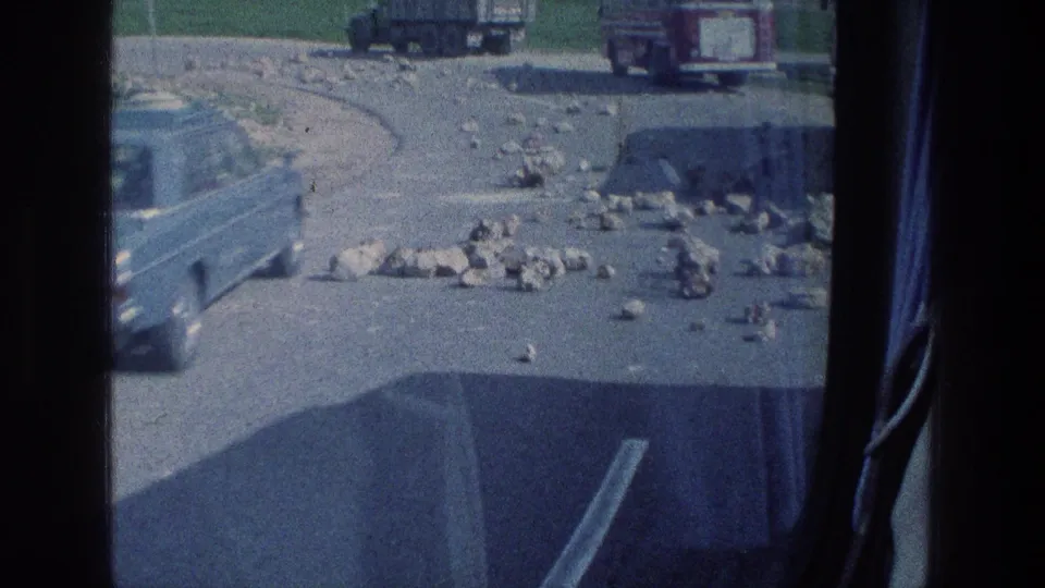 Passenger-side view in Israel from 1976, with scattered road stones, a pale vehicle blur, and distant trucks or cars beyond the window.
