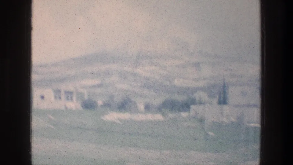 1970s 8mm home-movie frame from Israel showing a green field, hazy hills, and pale buildings in the distance.