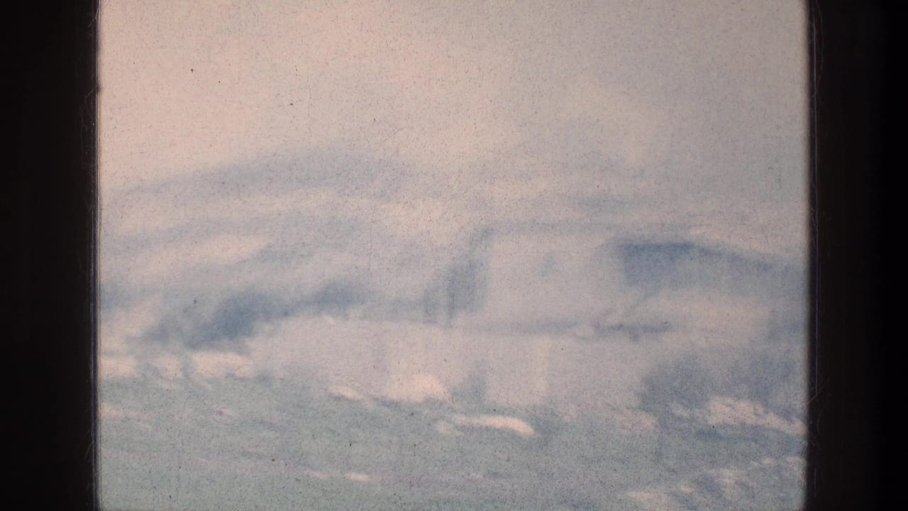 Blurred panning view of open green ground with low hills and pale structures beyond.