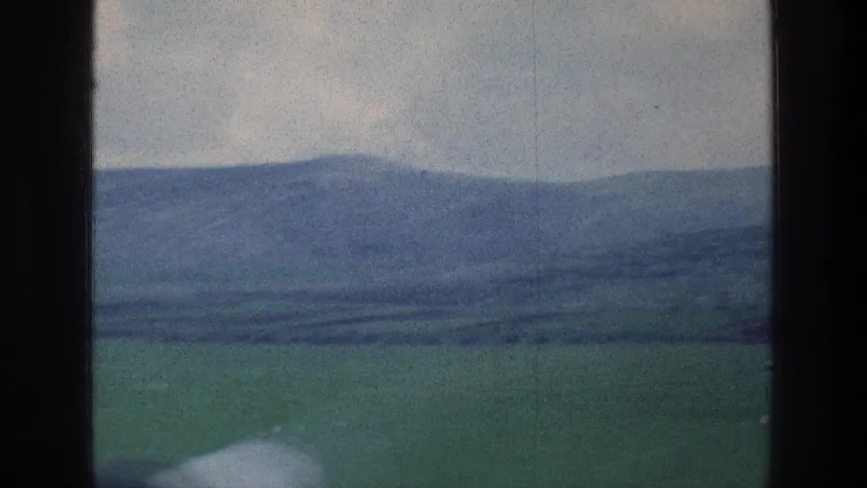 Muted 8mm frame showing open green countryside and hazy blue hills beyond a dark vertical window divider.