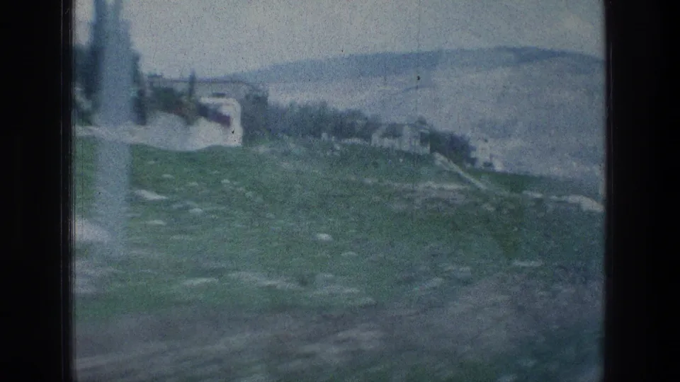 Vehicle-window view of a rocky green hillside with sparse trees and small structures under a hazy sky.