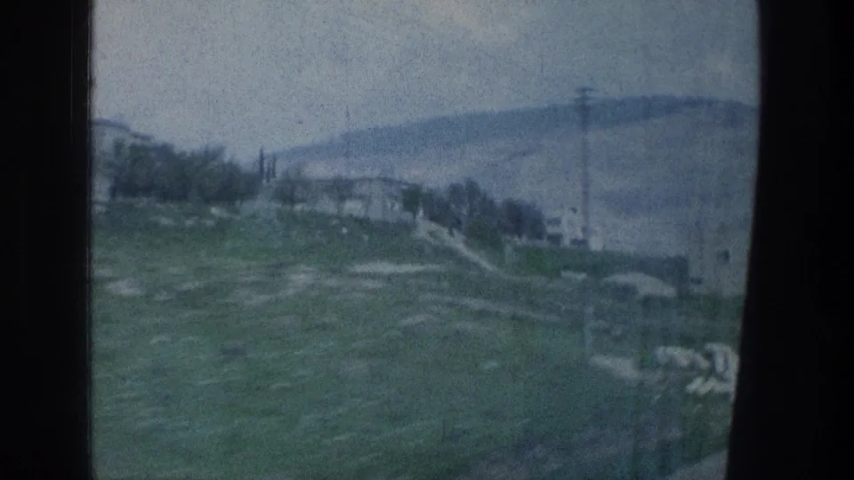 Soft 8mm travel frame with hillside buildings, trees, a tall utility pole, and distant mountains in hazy light.