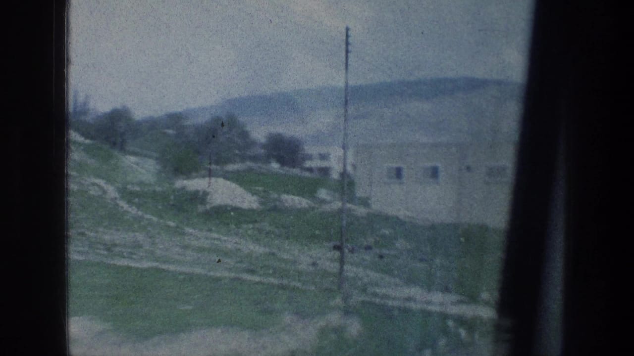 Passenger-view 8mm frame showing a hillside road, scattered trees, utility pole, and pale building in 1976 Israel.