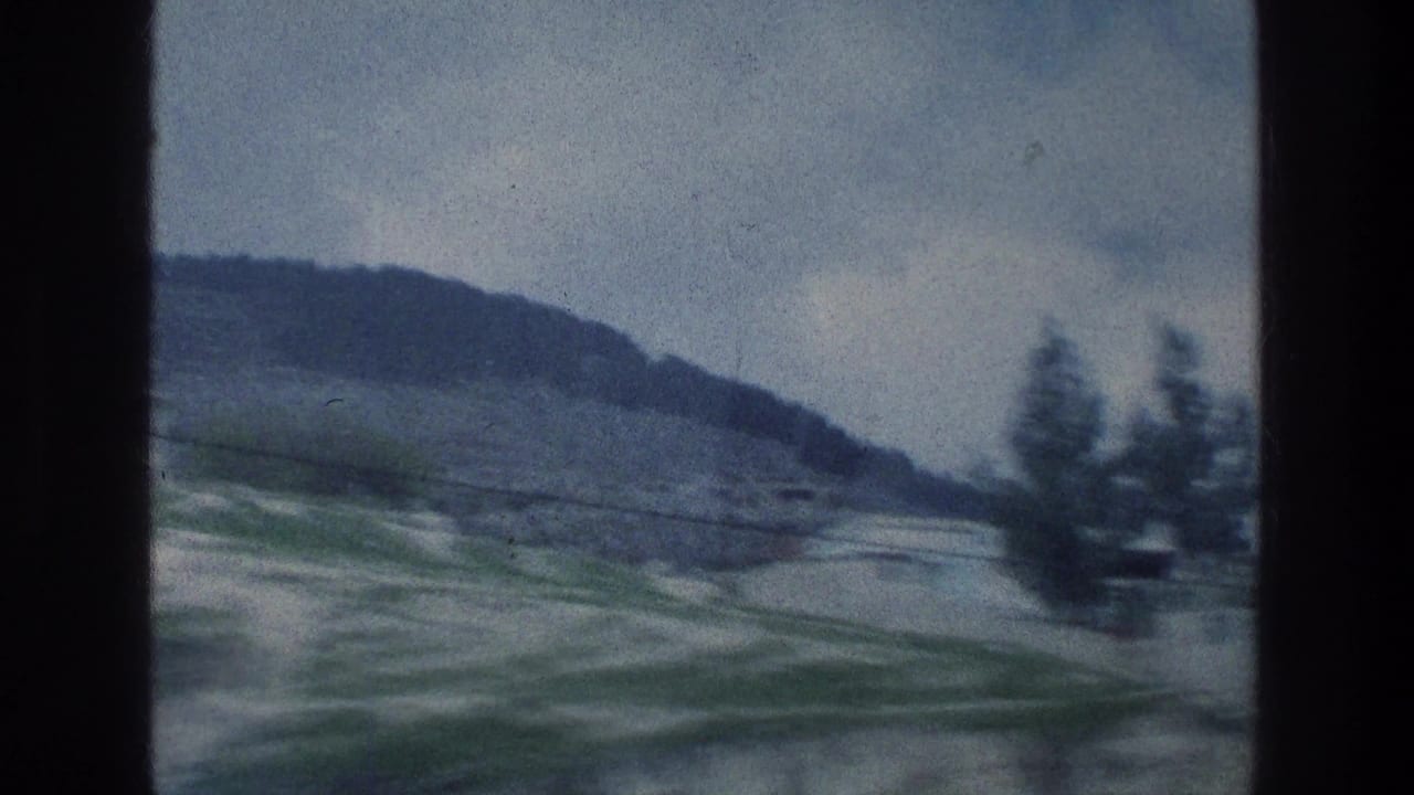 Curving road, green slopes, distant hillside, and dark vertical window frame.