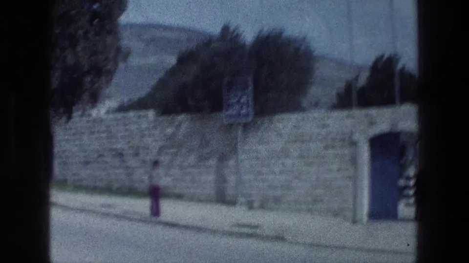 1976 8mm roadside view in Israel with a stone wall, small sign, blue door, trees, and hills beyond.