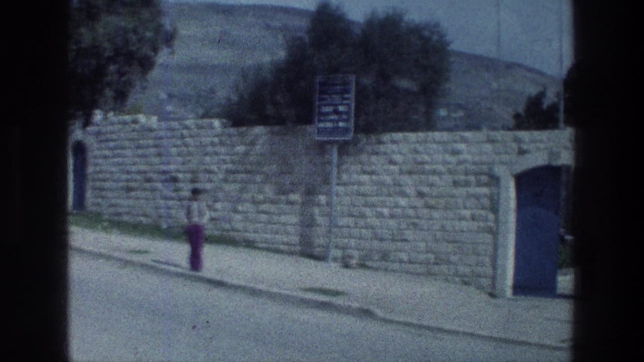1976 Israel home-movie frame showing a person walking along a road beside a stone wall, blue door, and sign.