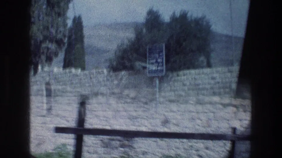 1976 Israel home-movie view through a window frame toward a stone wall, sign, trees, and hills beyond.