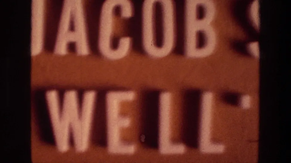 A tightly cropped brown sign shows raised white letters for Jacob's Well, with soft focus and dark vertical film edges.