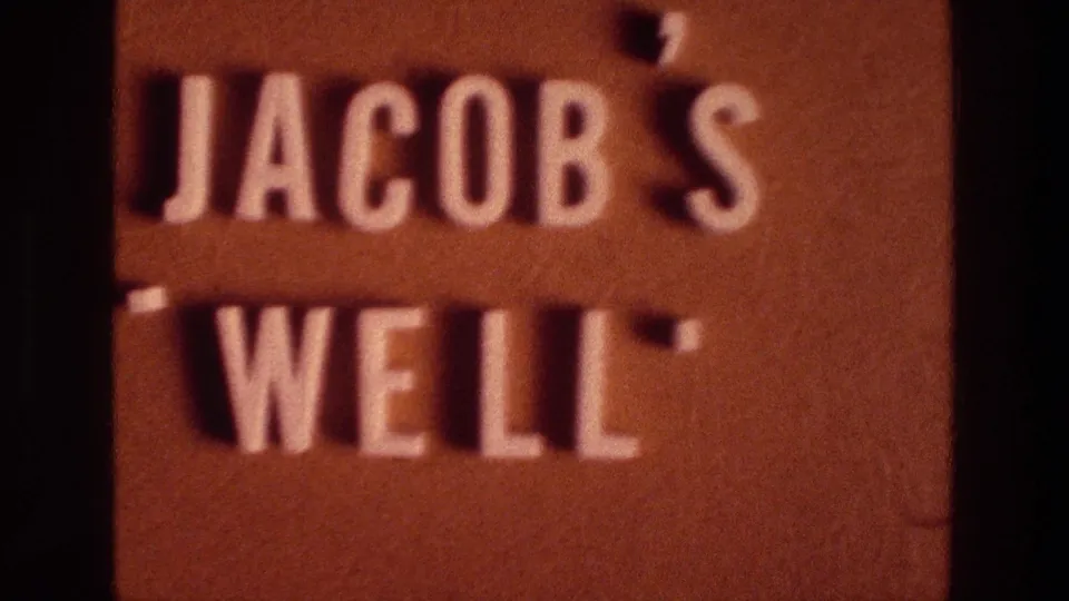 A later frame holds on the Jacob's Well sign, with the text centered lower in the image and a dark gate edge along the left side.
