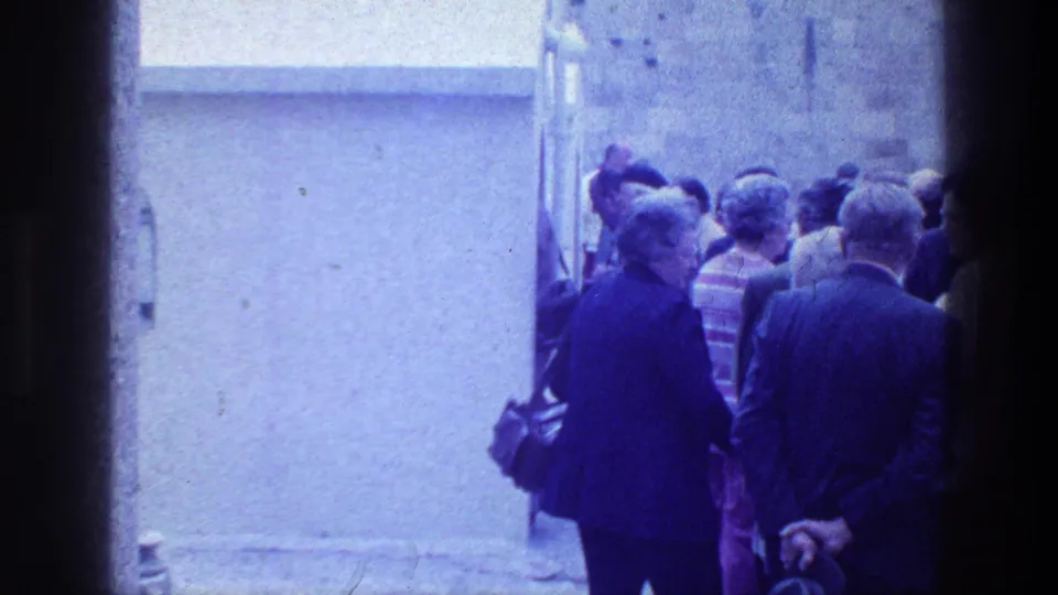 Adults in 1970s-style clothing gather in a narrow exterior space, viewed from behind on 8mm film.