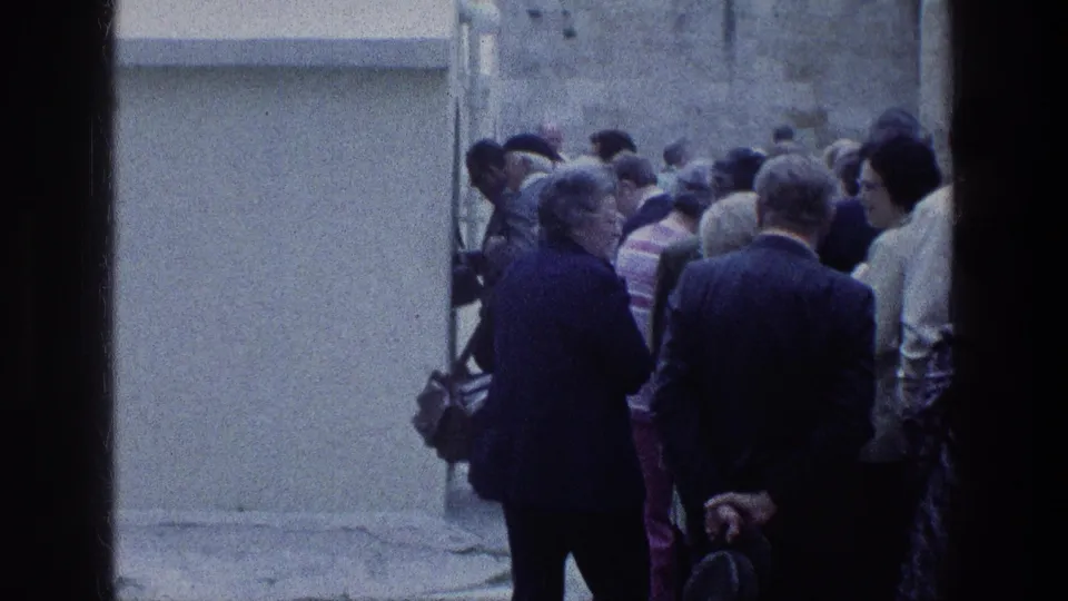 A small crowd of adults stands near a pale wall or entrance, with partial faces and period clothing visible.