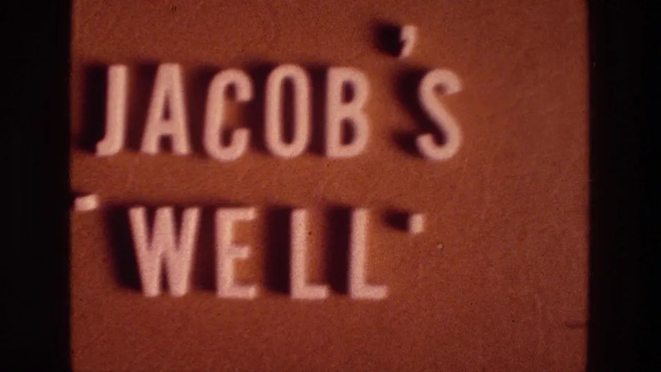 Late 1976 8mm frame holding on the Jacob's Well sign, with reddish-brown surface, raised white text, and soft film texture.