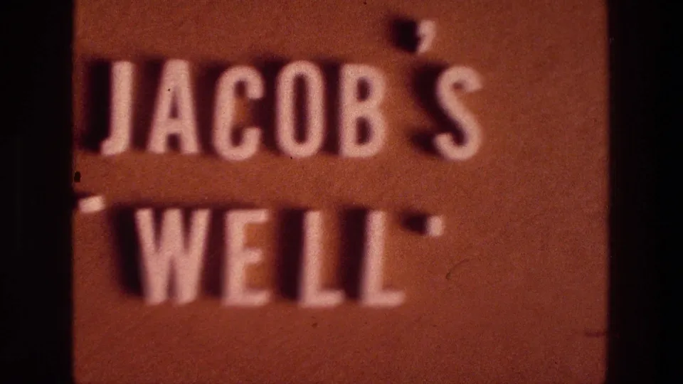 1976 8mm frame showing a reddish Jacob's Well sign partly obscured by the dark edge of a doorway.