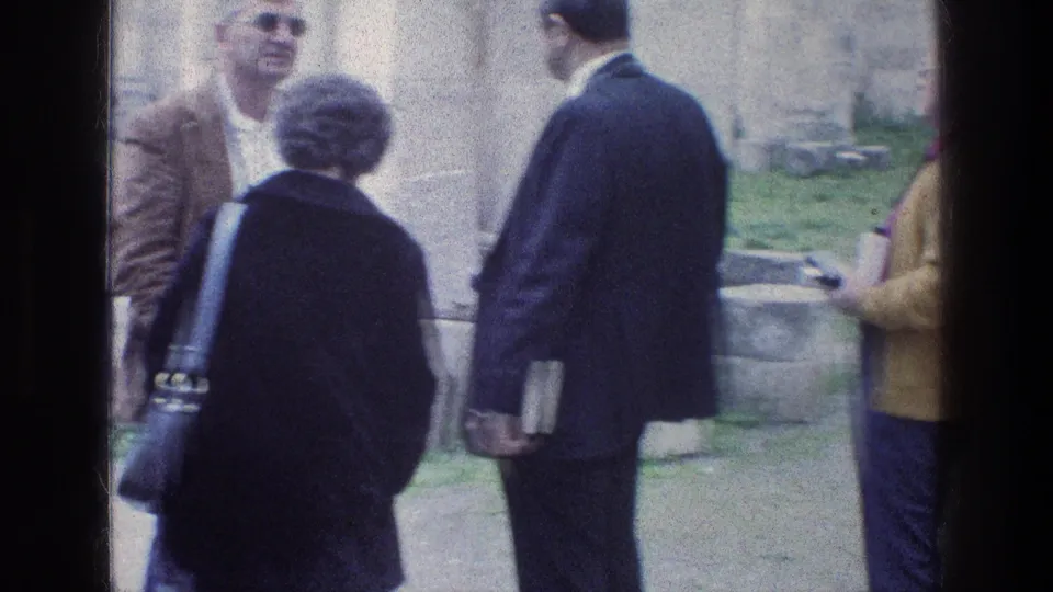 1976 8mm frame at Jacob's Well in Jerusalem showing adult visitors conversing near stone walls and a grassy edge.