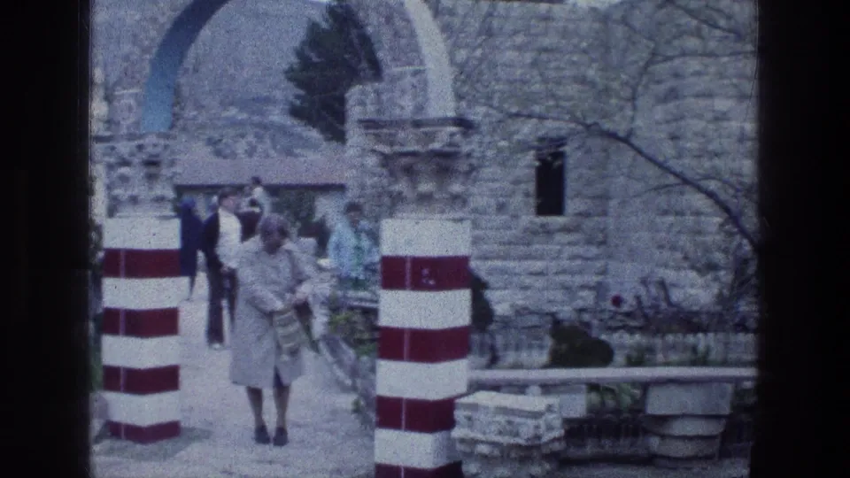 The woman in a grey coat walks beyond the red-and-white column at Jacobs Well, with stonework and benches visible behind.