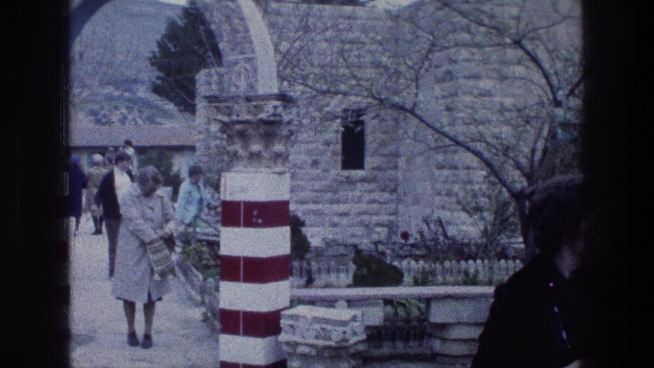 1976 8mm frame of visitors in coats moving through a stone courtyard at Jacob's Well in Jerusalem, with striped pillars and arches visible.