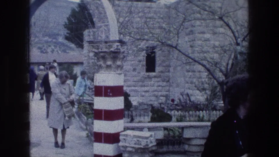 1976 8mm frame of visitors in coats moving through a stone courtyard at Jacob's Well in Jerusalem, with striped pillars and arches visible.