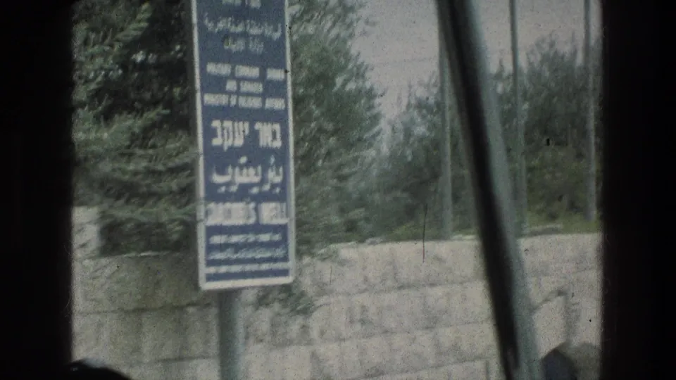 1976 8mm frame showing the Jacob's Well sign in Hebrew, Arabic, and English beside trees and a stone wall in Jerusalem.