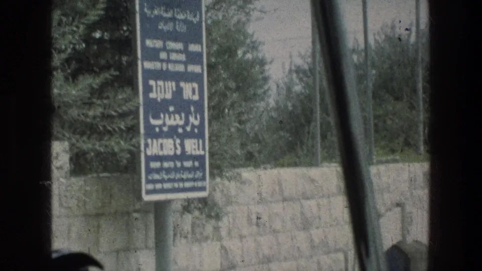 Handheld 8mm view of the Jacob's Well multilingual sign in Jerusalem, partially framed by dark film edges and nearby trees.