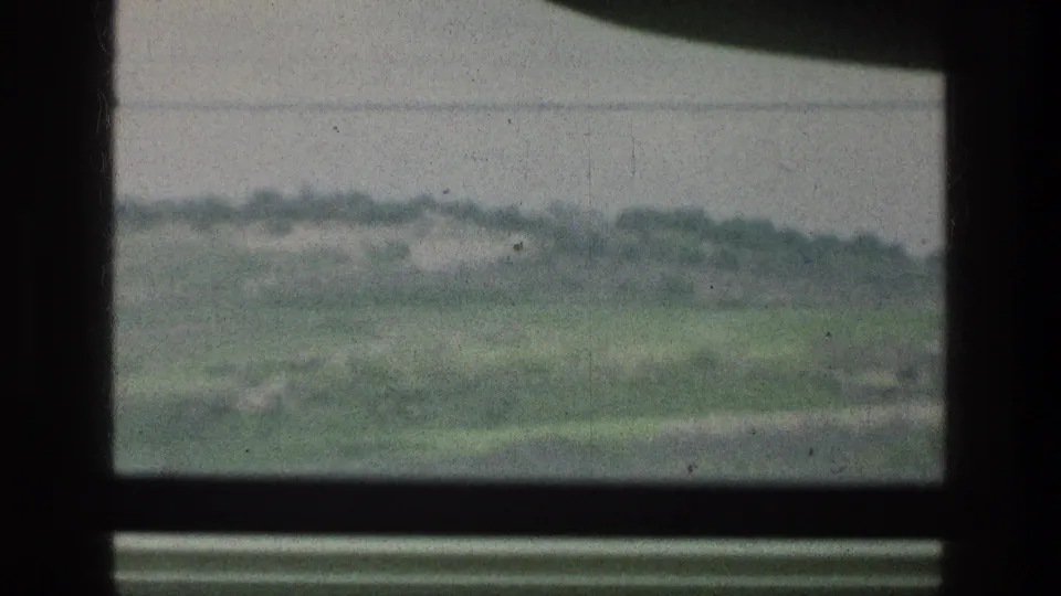 A 1976 8mm travel frame shows soft green slopes and scrubland through a dark lower window pane near the cataloged Jacobs Well area.