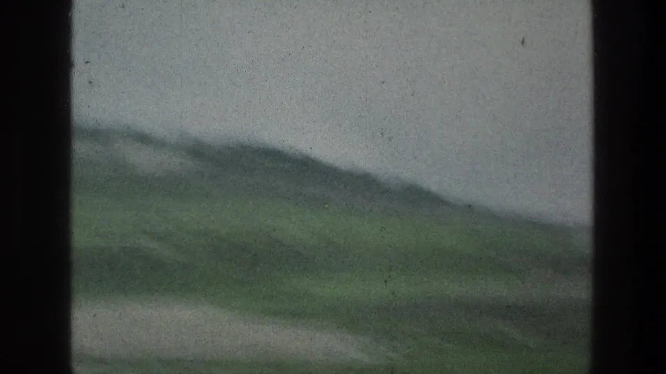 Soft 1976 8mm footage captures a passing hillside with green vegetation and a pale strip that may be water or road.