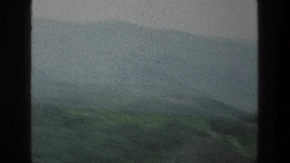 1976 home-movie frame of subdued green hills and distant slopes, with visible grain and dark frame edges.