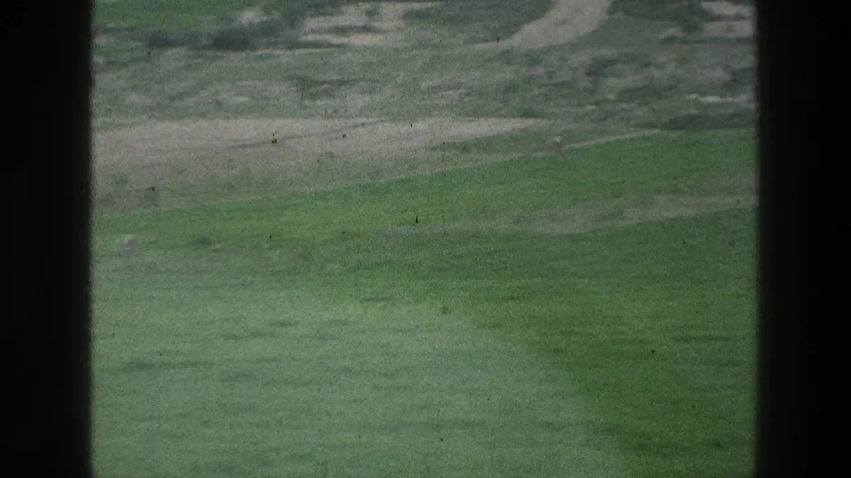 1976 8mm view of green fields near Jacobs Well metadata, framed by dark vertical edges with faint distant figures.