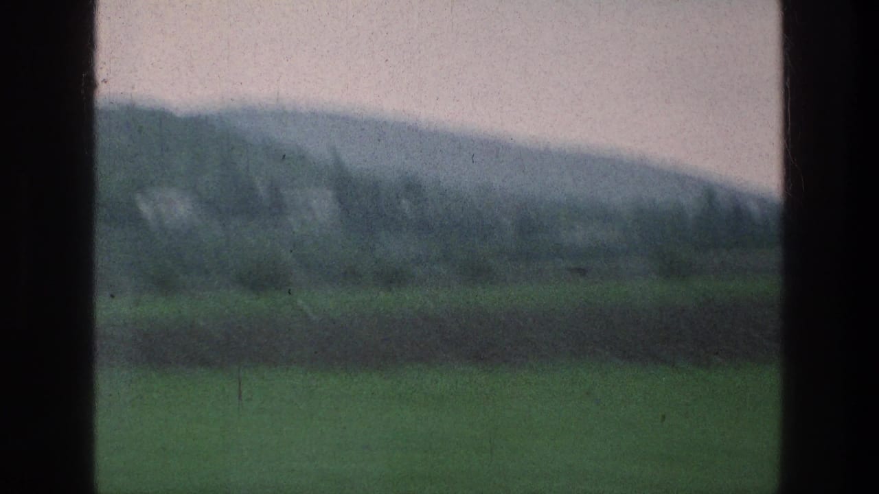 Rural 1976 train-window view with green fields, darker cultivated strips, and low hills under a pale sky.