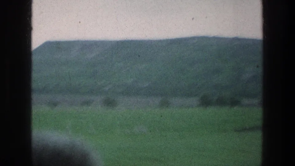 Soft 8mm frame of green farmland and a hazy hillside, seen from a moving train in the listed 1976 Jerusalem-area travel footage.