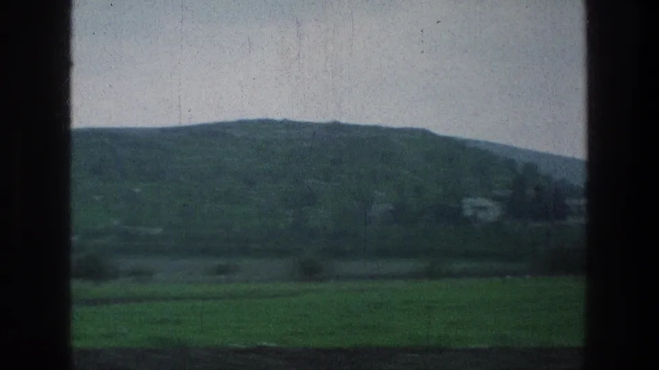 1976 8mm countryside view with layered fields, low hills, and small distant structures seen through dark train-window framing.