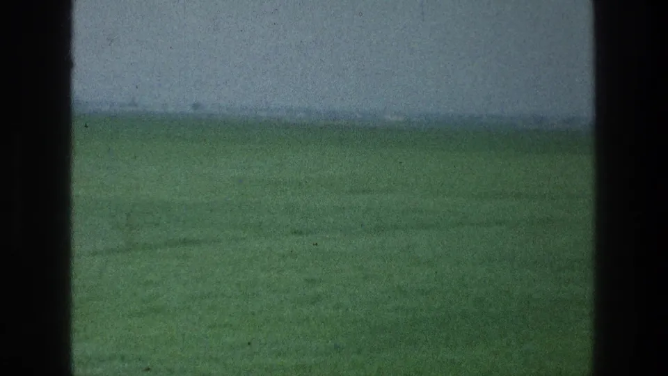 1976 8mm frame from the Jacobs Well Jerusalem Israel metadata set showing a green field under a hazy sky, framed by dark foreground edges.