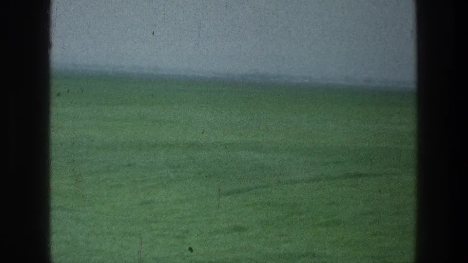 1976 8mm landscape frame showing a broad green field and muted horizon, partially obscured by dark vertical foreground bands.