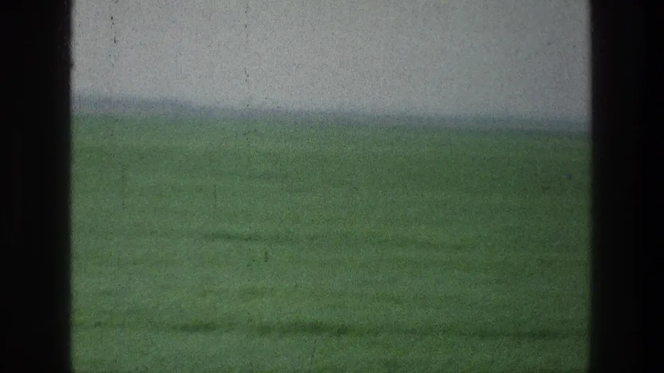 1976 8mm frame of a green rural field viewed through dark window framing, with a soft hazy horizon.