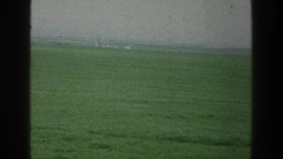A soft 8mm view of a green field through window framing, with muted color and visible film wear.