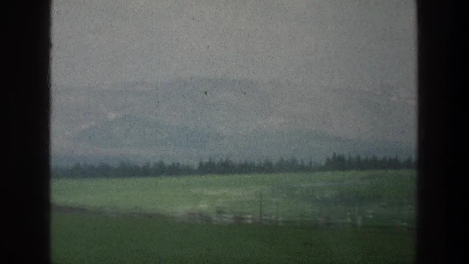A 1976 8mm travel-view frame shows green fields, a distant wooded ridge, hazy hills, and dark framing from a window or film edge.