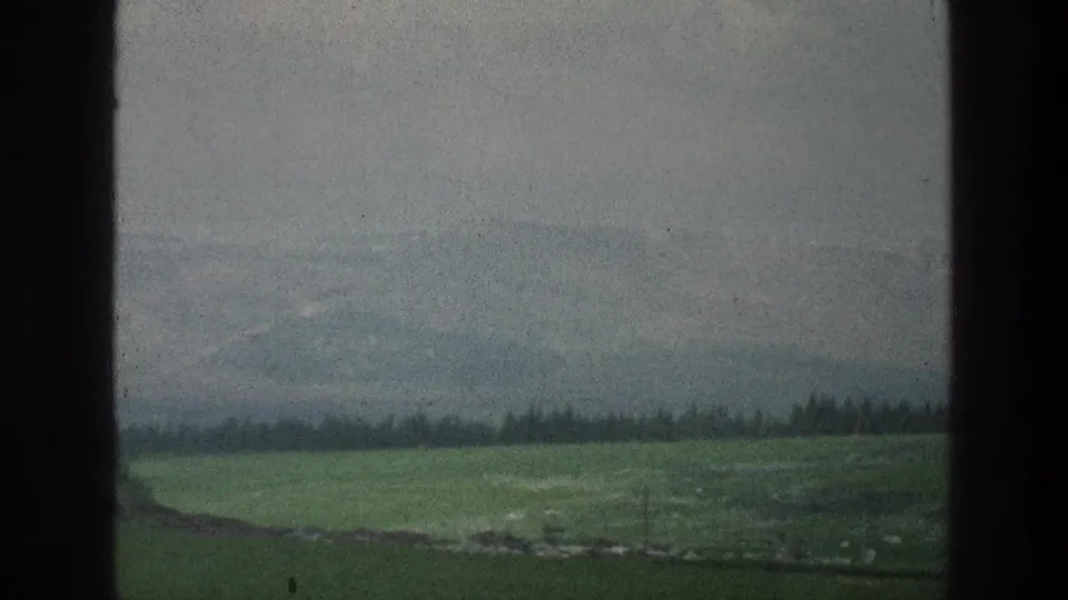 Muted 8mm landscape from the stated 1976 Jerusalem, Israel footage, showing green fields, distant hills, and a dark window-like edge.
