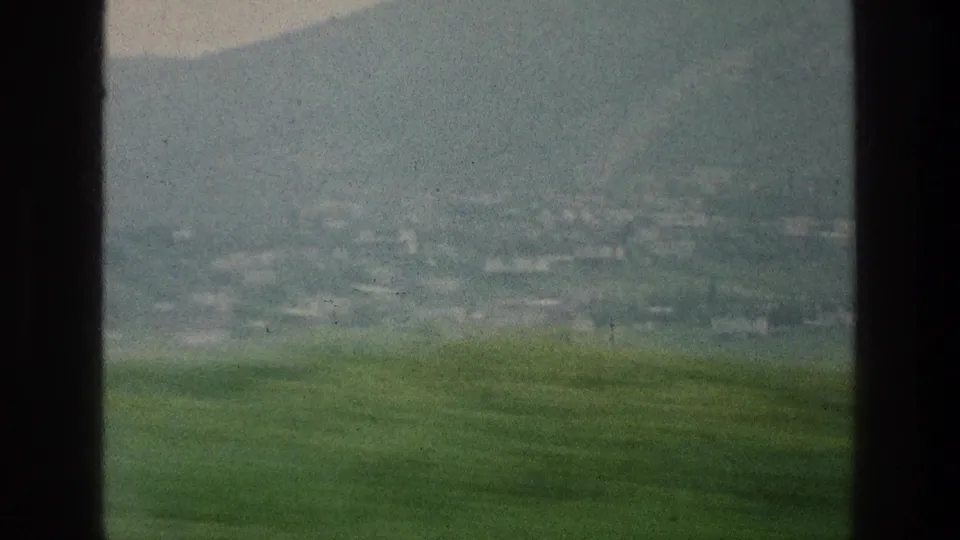 1976 Israel travel view with green foreground, hazy mountain slope, and faint distant settlement-like structures.