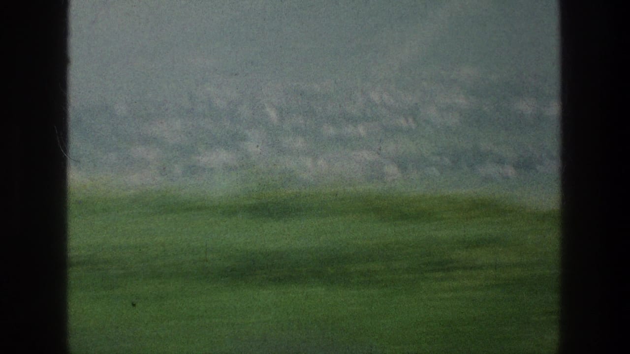 Hazy green field and distant hillside seen through dark vehicle window frames.
