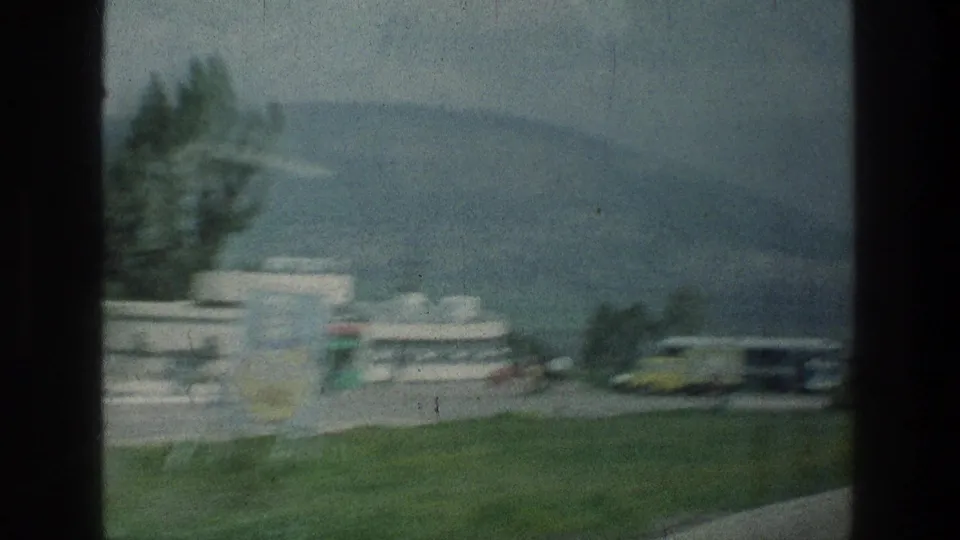 Vehicle-shot 1976 8mm view near Jacobs Well Jerusalem, showing a roadside sign, low structures, parked cars, trees and hills.