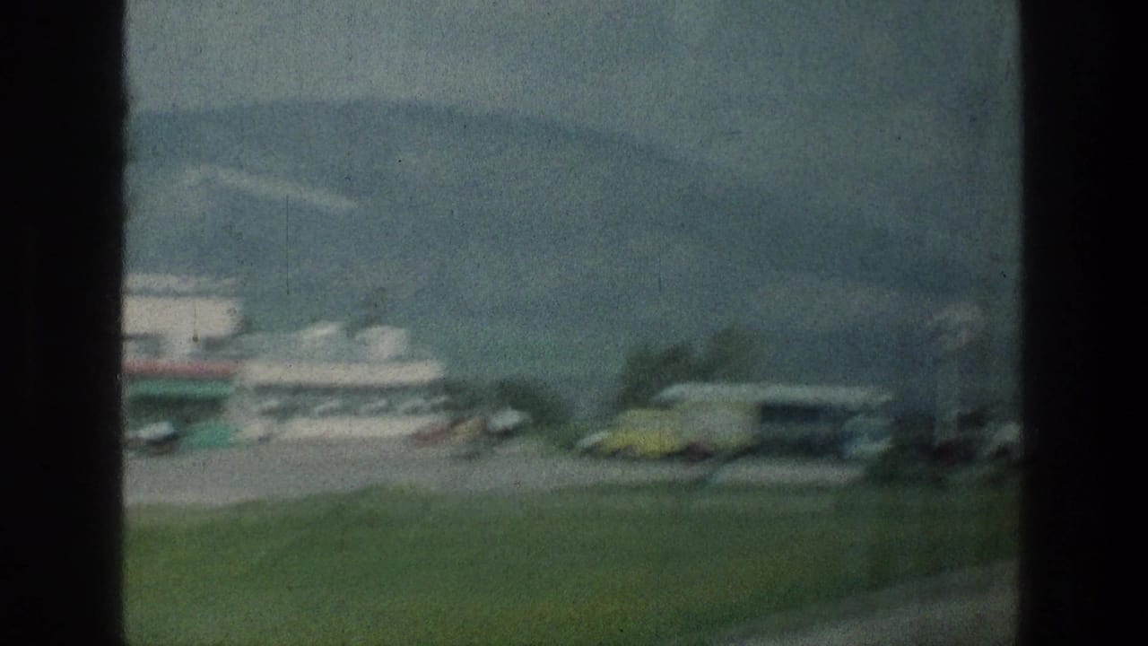 1976 8mm roadside view near the stated Jerusalem-area location, showing a gas station area, parked vehicles, grass, and distant mountains.