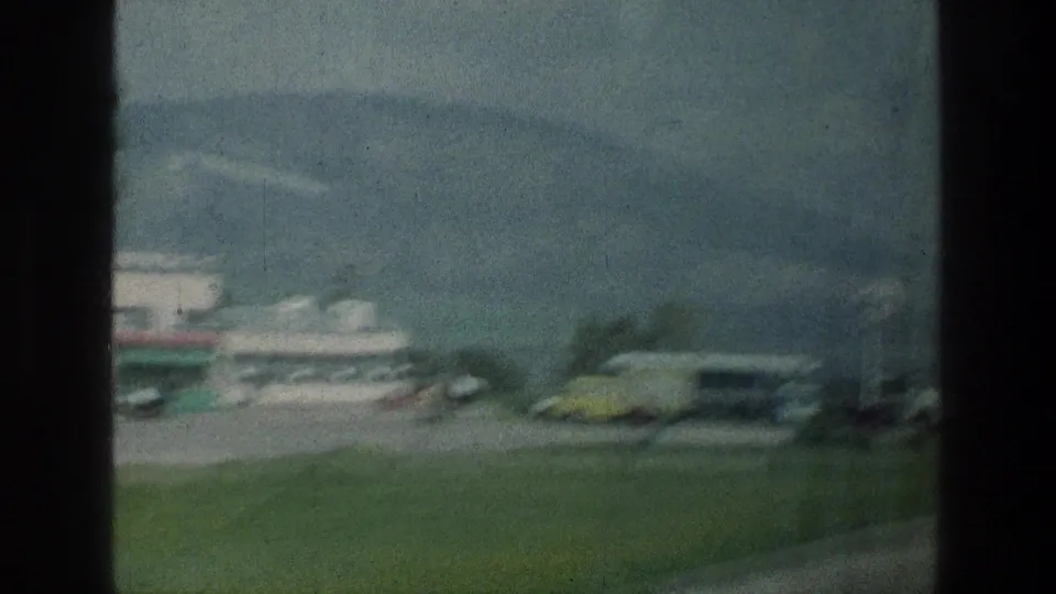1976 8mm roadside view near the stated Jerusalem-area location, showing a gas station area, parked vehicles, grass, and distant mountains.
