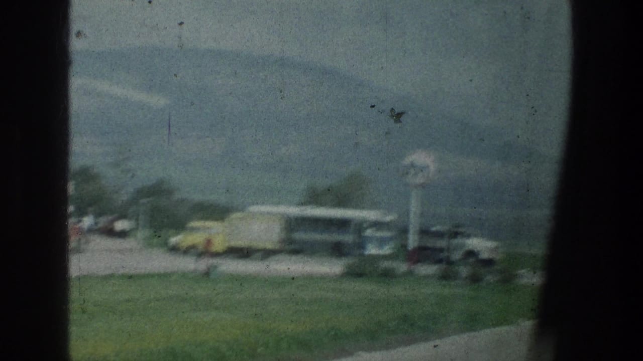 Gas station sign, bus, trucks, low building, grass, and mountain backdrop seen through film gate edges.