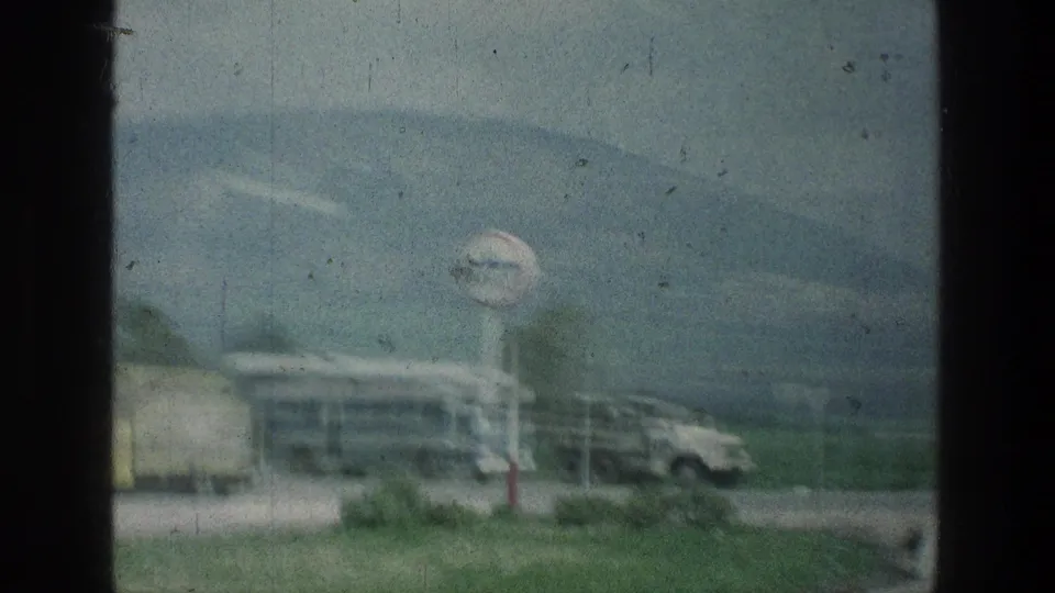 1976 8mm frame showing a roadside sign, bus, truck or trailer, car, grass, and mountain backdrop at the stated Jerusalem-area location.