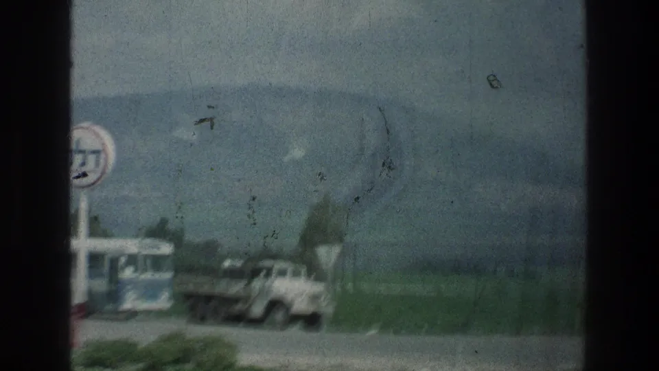 1976 8mm frame with a military-style truck, roadside sign, bus, open grass, and distant mountains near the stated Jerusalem-area location.