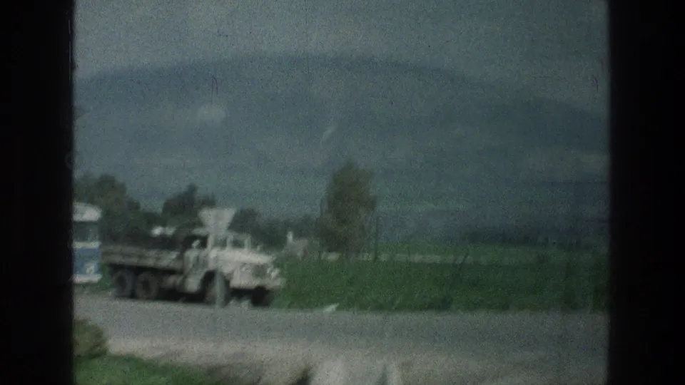 1976 8mm view near the stated Jerusalem-area location, showing a white roadside truck, green fields, trees, and a distant mountain through dark frame edges.