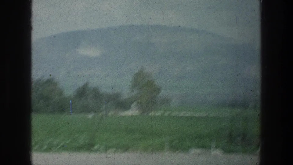 Grainy 1976 home-movie frame showing green fields, blurred trees, roadside foreground, and a hazy mountain under pale sky.