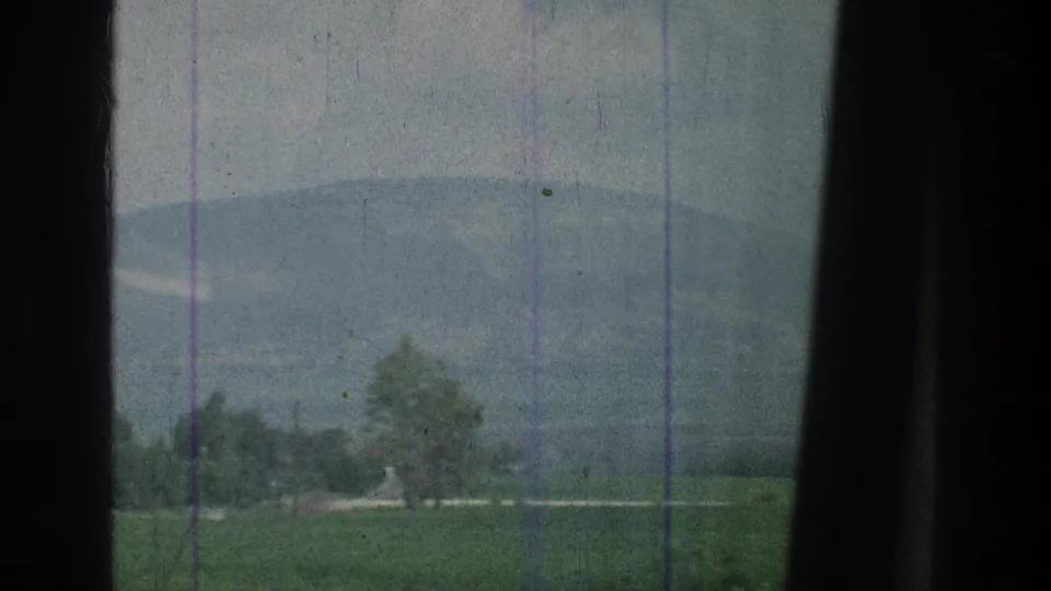 Final 8mm frame of the passing Jerusalem-area landscape, with fields, small trees, a distant mountain, vertical scratches, and dark edge framing.