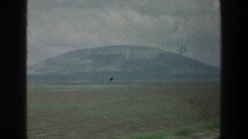 A hazy landscape view through a window shows a broad field, rounded hill, and overcast sky in 1976 8mm footage.